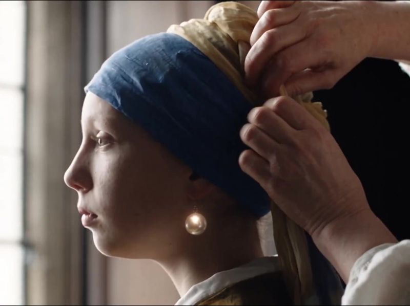 Still uit de AI-film: een vrouw wikkelt een gele doek over de blauwe band op het hoofd van een meisje met een parel in haar oor